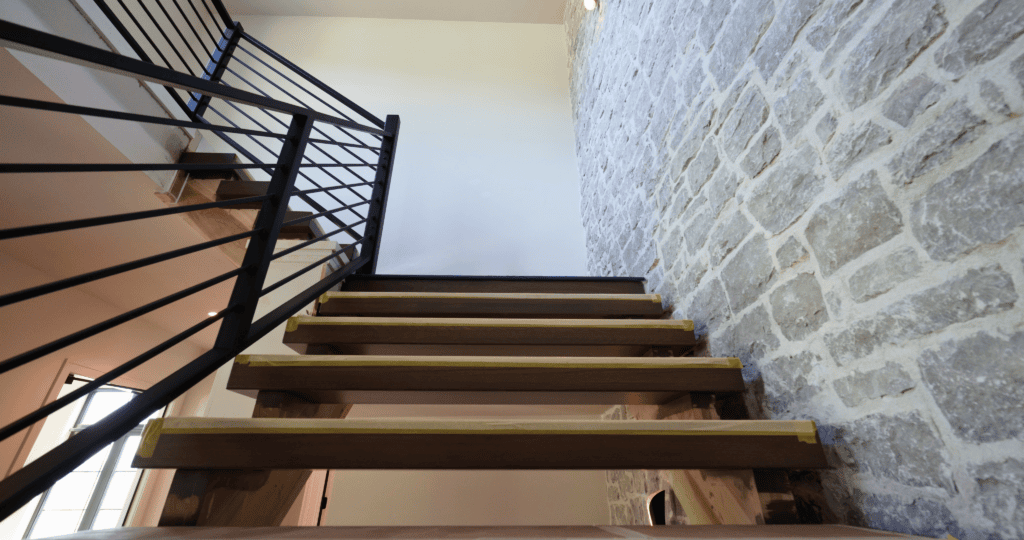 Floating wood stair treads with a black metal rail set against a natural stone wall inside The Ironwood