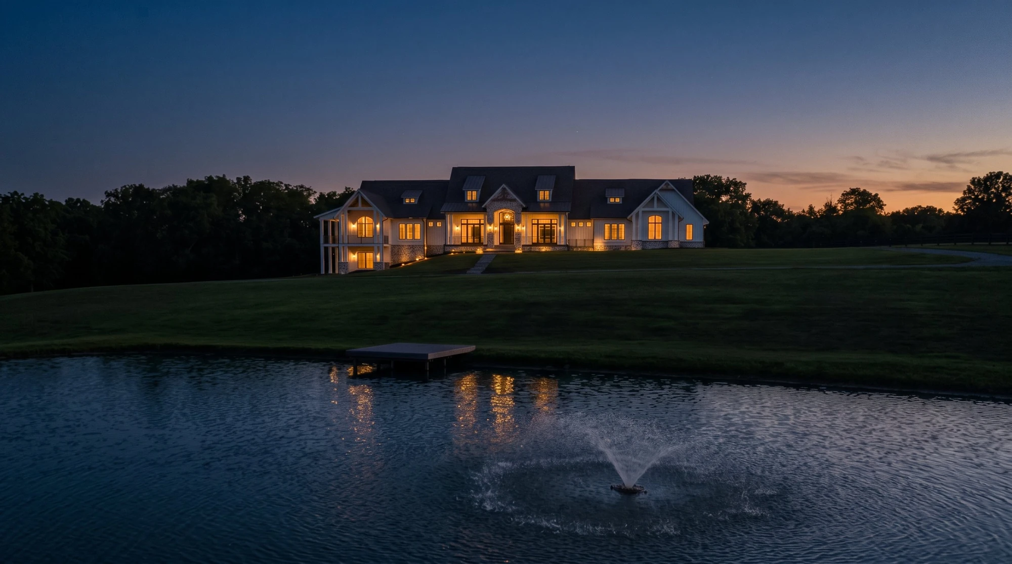 Justin Doyle Homes custom ranch with a walkout lower level at dusk, every window lit, seen across a pond with a fountain and a small dock