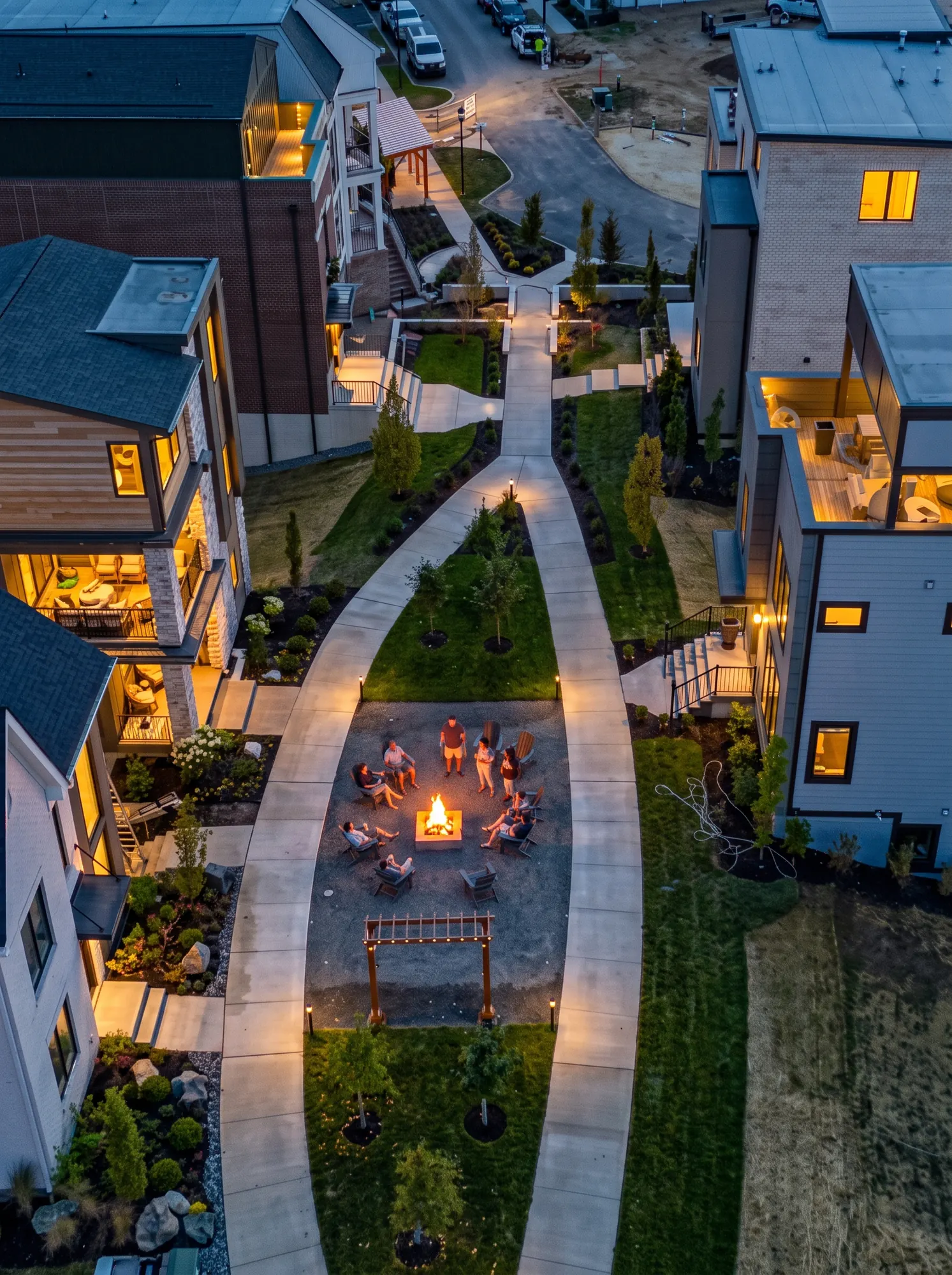 Aerial view at dusk of people in Adirondack chairs around a fire pit in a Foundry Park green space, between lit walkways and three- and four-story homes