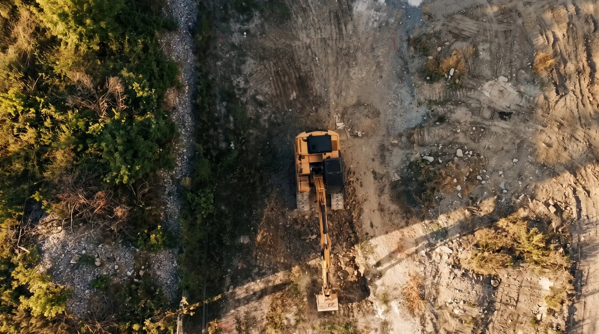 Overhead drone view of an excavator working a cleared homesite at golden hour, with track marks in the soil and brush along one edge